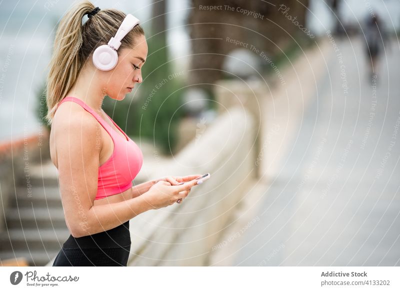 Sportive woman in headphones using social media on smartphone sportswoman sportswear listen music embankment sea gadget device internet online cellphone