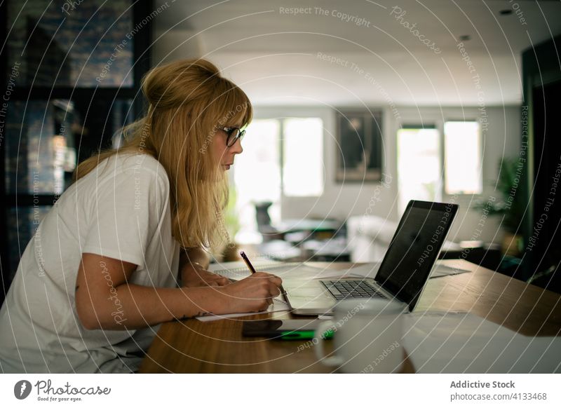 Confident female freelancer taking notes and using laptop take note work woman paper telework serious entrepreneur project confident netbook modern apartment