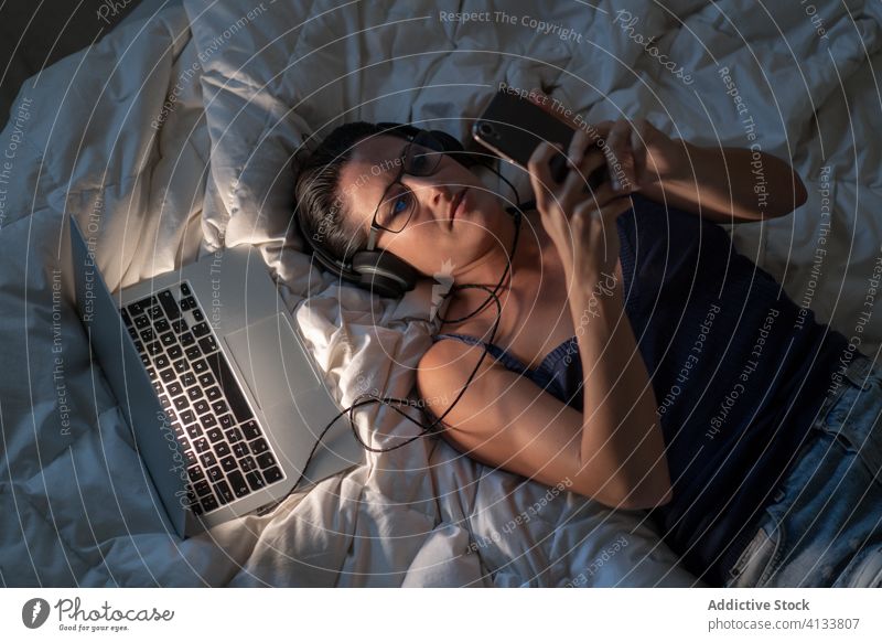 Calm woman listening to music in headphones on bed relax smartphone enjoy using song female browsing blanket lying down tank top device technology laptop gadget