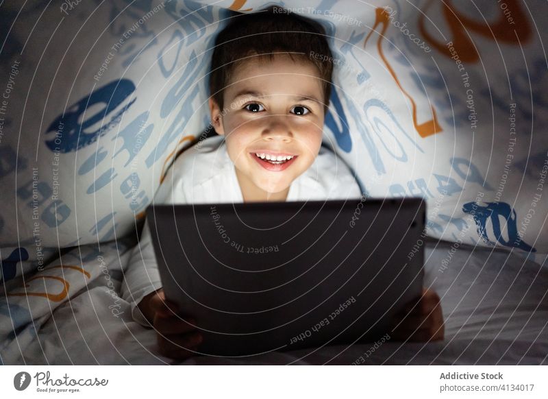 Delighted boy watching cartoon on tablet kid using blanket night cheerful pajama home adorable device gadget child internet browsing cozy rest cute relax online