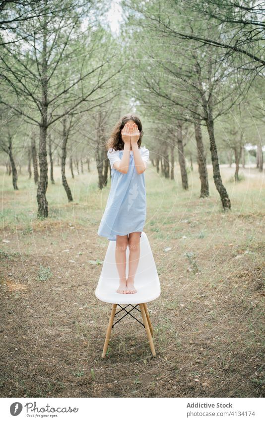 Unrecognizable girl standing on chair in alley between trees cover face park peaceful harmony barefoot hide concept casual dress child idyllic trunk youth