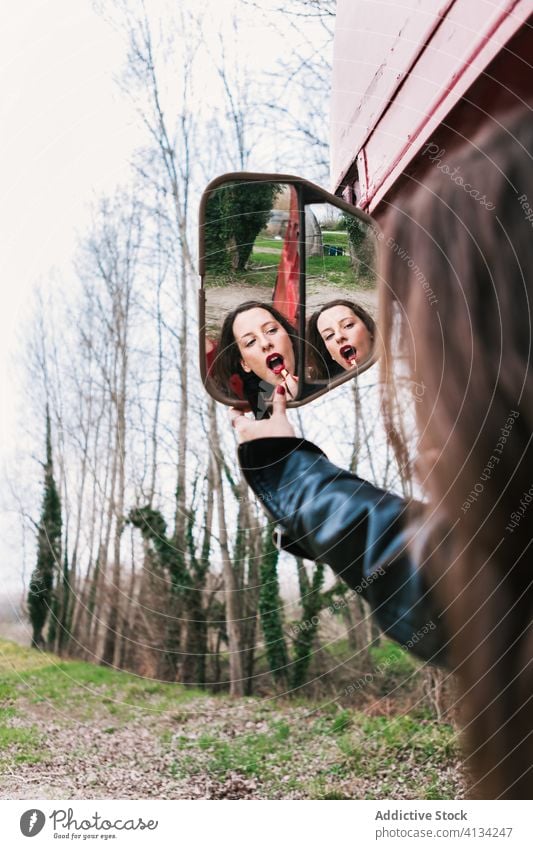 Woman applying lipstick and looking in side mirror of bus woman makeup charming red lips leather jacket put on female forest side view mirror wood nature