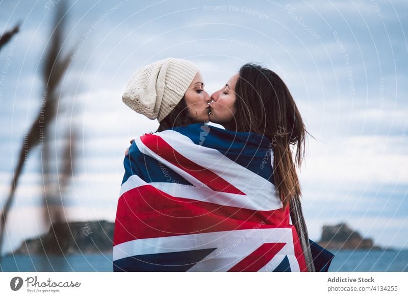 Kissing young lesbian couple with Great Britain flag great britain seashore cloudy sky kiss hug wrap women eyes closed hat lifestyle embrace gay bonding lgbt