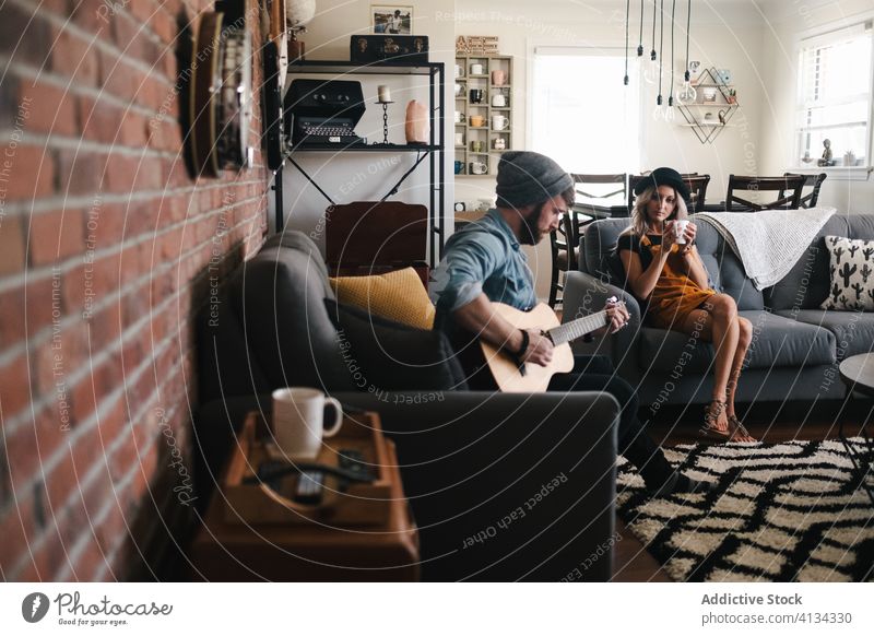 Musician playing guitar sitting with girlfriend on sofa at home couple music free time comfort living room friendship together relationship legs crossed