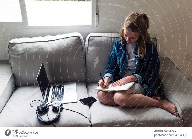 Millennial woman sitting on sofa with gadgets and writing in notebook write home young take note diary concentrate serious female casual focus device couch