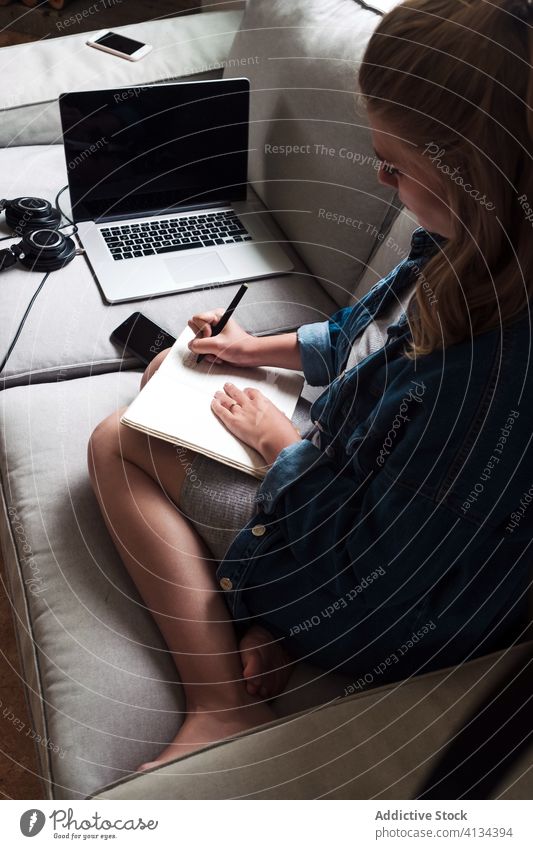 Millennial woman sitting on sofa with gadgets and writing in notebook write home young take note diary concentrate serious female casual focus device couch