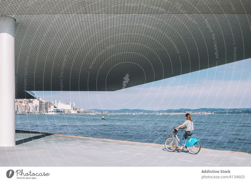 Woman riding bicycle along seafront ride city woman modern building architecture sunny bike female transport waterfront summer street contemporary style hobby
