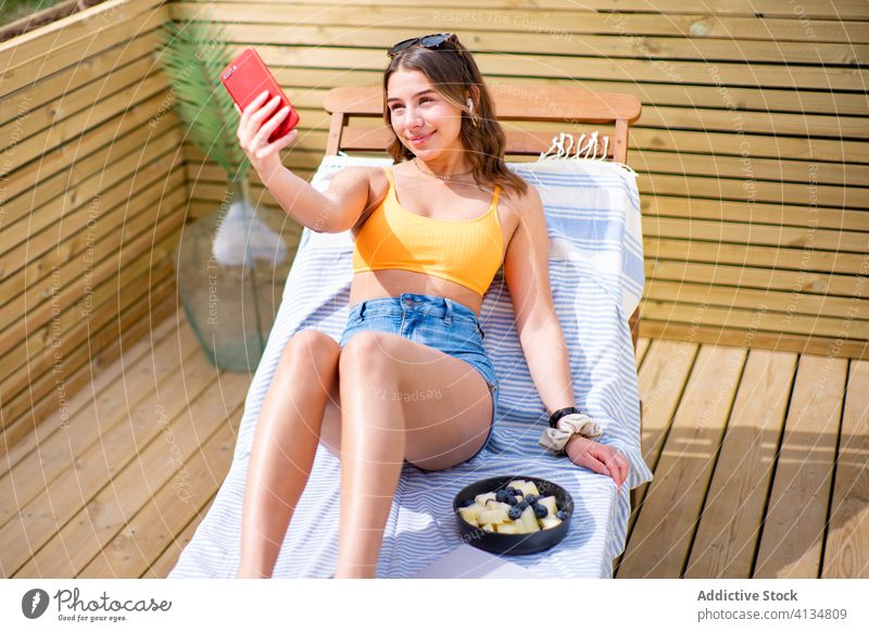 Charming woman taking selfie on smartphone on deck chair deckchair summer positive sunny camera relax using female charming shorts bra lying cellphone gadget