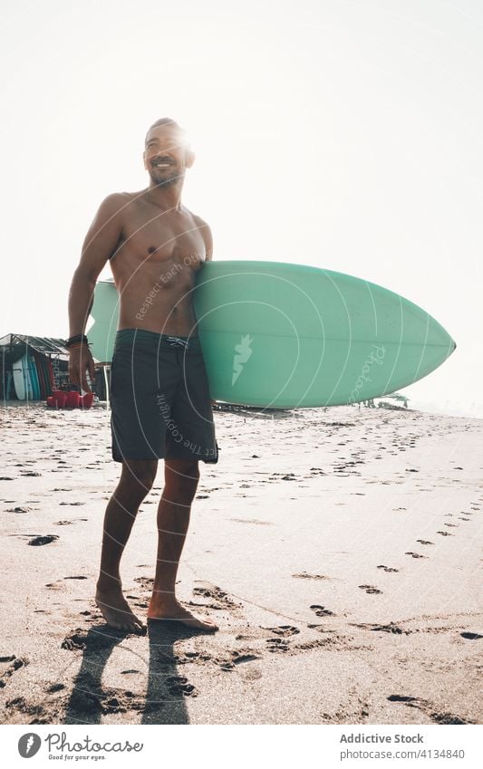 Cheerful surfer with surfboard standing on beach man happy seafront active ocean coast smile shirtless ethnic male lifestyle sand summer shore vacation water