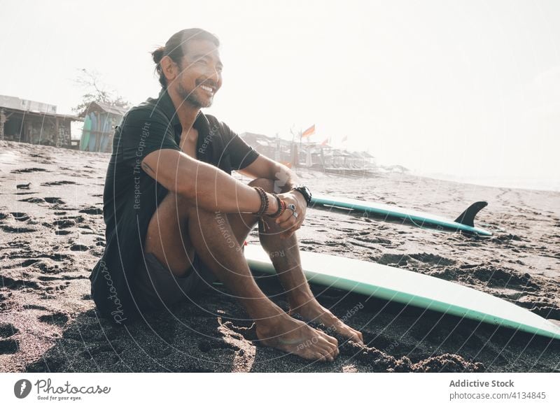 Happy ethnic man with surfboard sitting on beach happy positive rest sea enjoy sand summer young hispanic male lifestyle vacation coast ocean shore journey glad