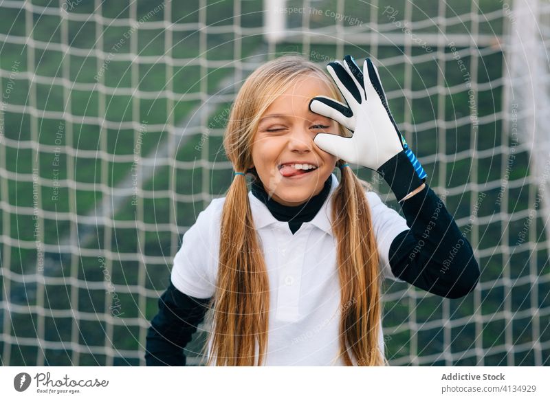 Cheerful preteen girl goalkeeper teasing rival on football field at sports stadium soccer glove make face training playful cheerful net female uniform