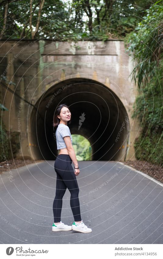 Slim Asian woman standing on asphalt road near tunnel tree plant sportswear weekend recreation toothy smile positive holiday happy enjoy summer content cheerful
