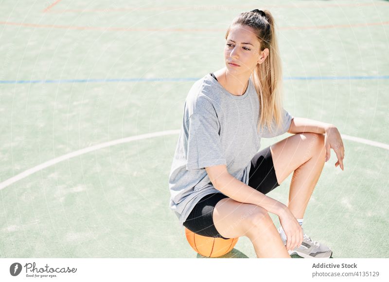 Sporty woman sitting on basketball ball on court confident sport rest serious young sportswoman athlete sportswear training activity game player workout break