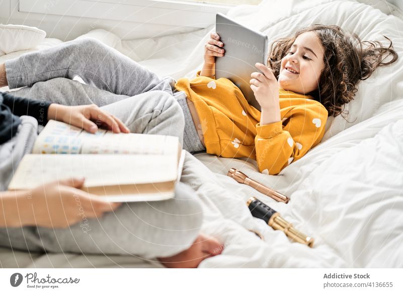 Calm siblings relaxing on bed with book and device tablet girl boy read watch using weekend children kid childhood lounge chill cozy calm rest interesting