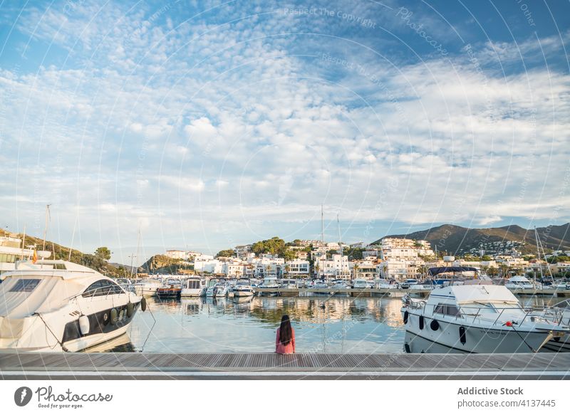 Anonymous woman sitting in port of coastal town pier travel vessel picturesque coastline sea seashore female llanca spain water reflection cloudy trip vacation