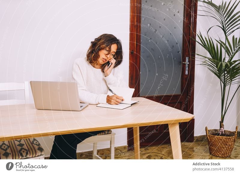 Busy woman working remotely from home laptop phone call take note busy distance female modern casual entrepreneur table conversation project freelance gadget