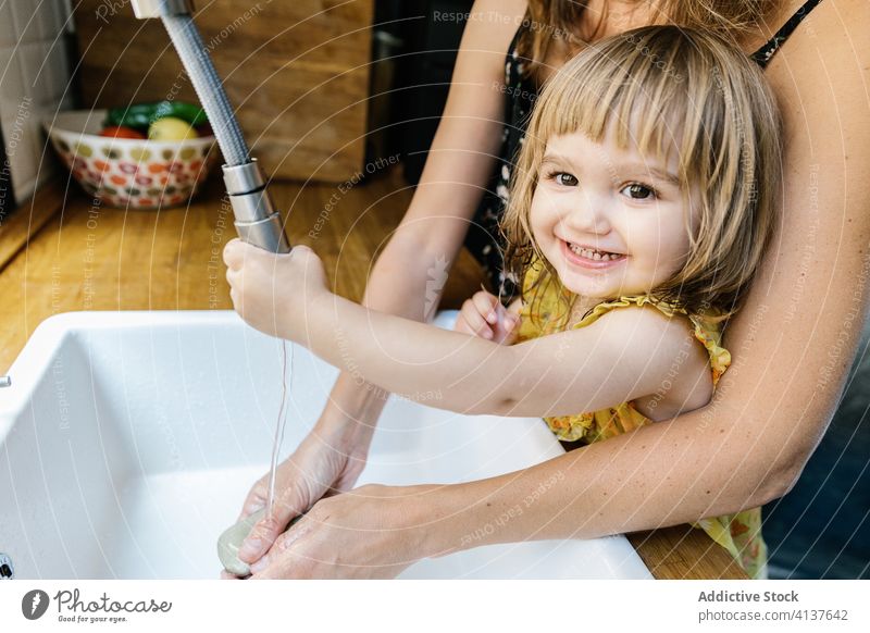 Cheerful girl washing hands with mother daughter soap smile teach kitchen home sink tap water hug embrace little lifestyle childhood domestic kid together