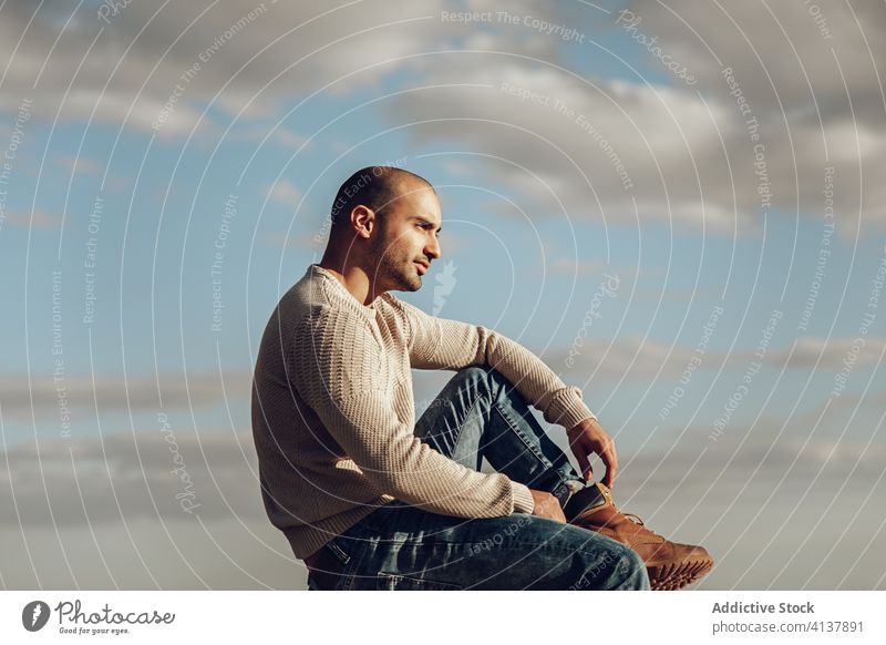 Hipster man sitting against cloudy sky pensive hipster style trendy modern street style thoughtful contemplate clouds sunny young unshaven guy jeans boot casual