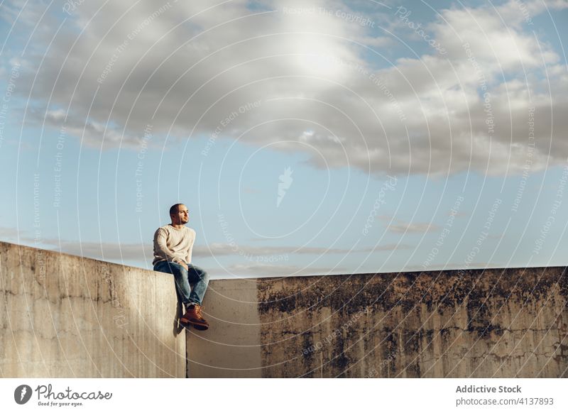 Young man sitting on concrete construction on street fence stone shabby urban alone hipster modern sky clouds sunny guy casual male confident contemporary