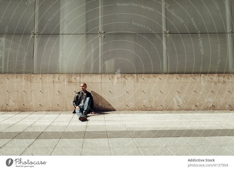 Ethnic man in trendy outfit sitting on street hipster young style building contemporary portrait modern guy casual ripped jeans beard urban concrete wall