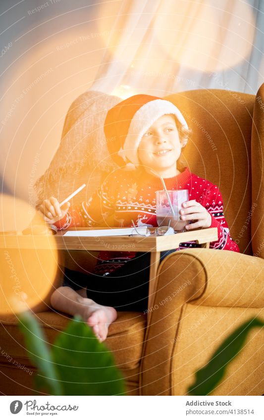 Happy boy in Christmas wear sitting in cozy armchair santa hat christmas holiday pencil happy comfort domestic sweater glad cheerful comfortable bokeh positive