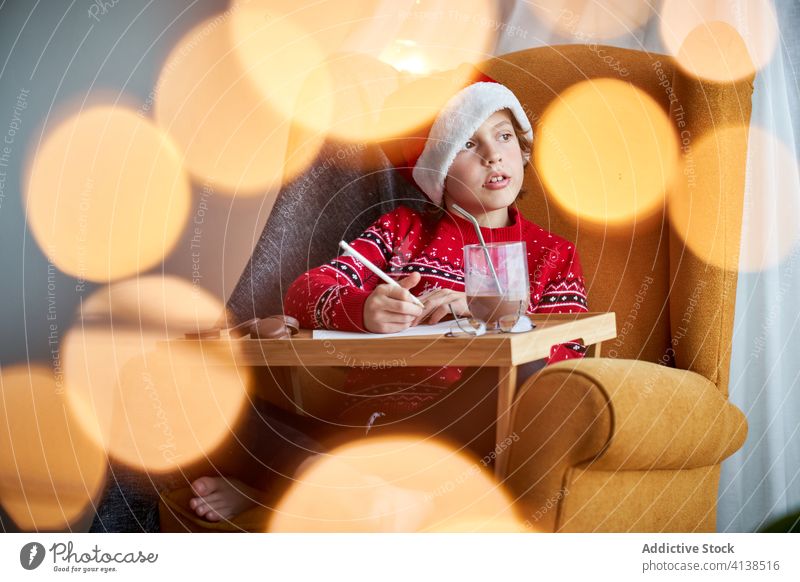 Boy in Christmas wear sitting in cozy armchair boy santa hat christmas holiday pencil happy comfort domestic sweater glad cheerful comfortable bokeh positive