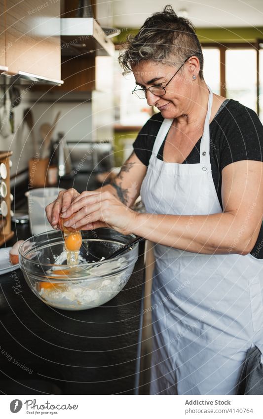 Mature female cooking in kitchen woman prepare dough housewife pastry add egg ingredient flour bowl mature middle age table culinary home recipe apron homemade