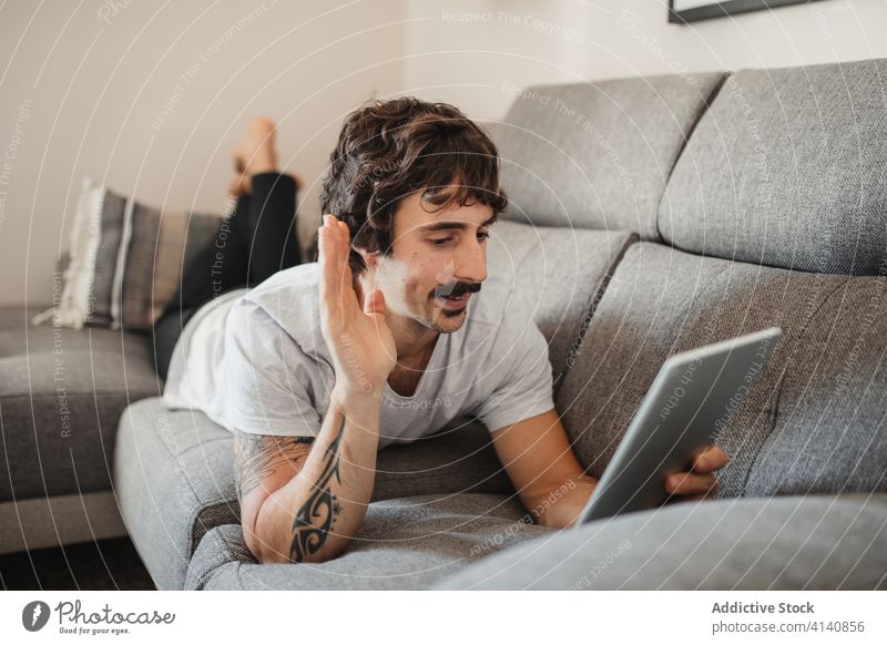 Content man talking on video chat via tablet video call home wave hand greeting gesture speak male relax weekend couch lying casual gadget device sofa modern
