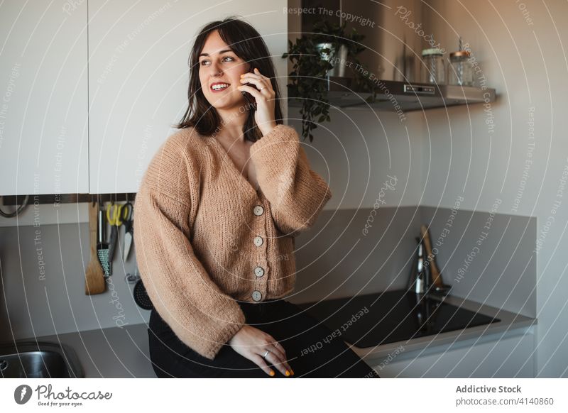 Cheerful woman talking on smartphone at home conversation phone call using female trendy sweater kitchen speak cheerful sit device gadget smile joy communicate