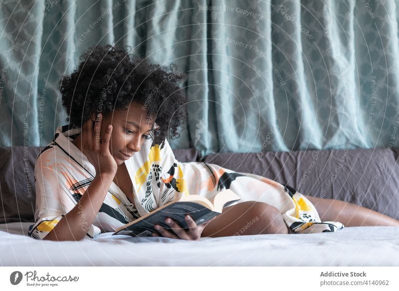 Focused ethnic woman reading book at home enjoy novel rest comfort bed content female black african american lying silk robe relax bedroom peaceful cozy calm