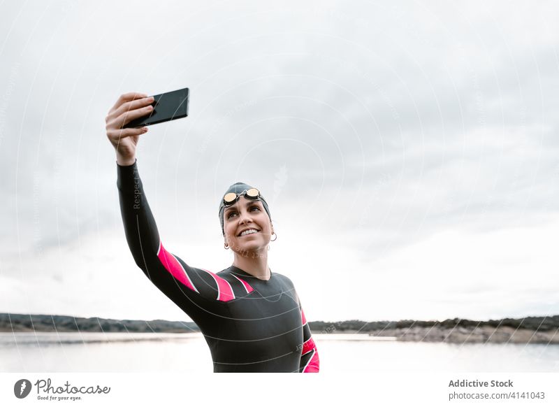 Cheerful female diver taking selfie on calm shore woman happy wetsuit social media lake sport cheerful nature inspiration goggles smartphone cap sunset barefoot
