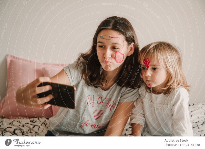 Sisters with painted faces taking selfie on smartphone sister entertain sibling girl play using little teenage home having fun cellphone photo bed kid camera