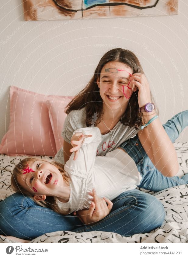 Sisters with painted faces cuddling on bed sister laugh having fun weekend enjoy girl teenage little creative cheerful child kid casual childhood together home