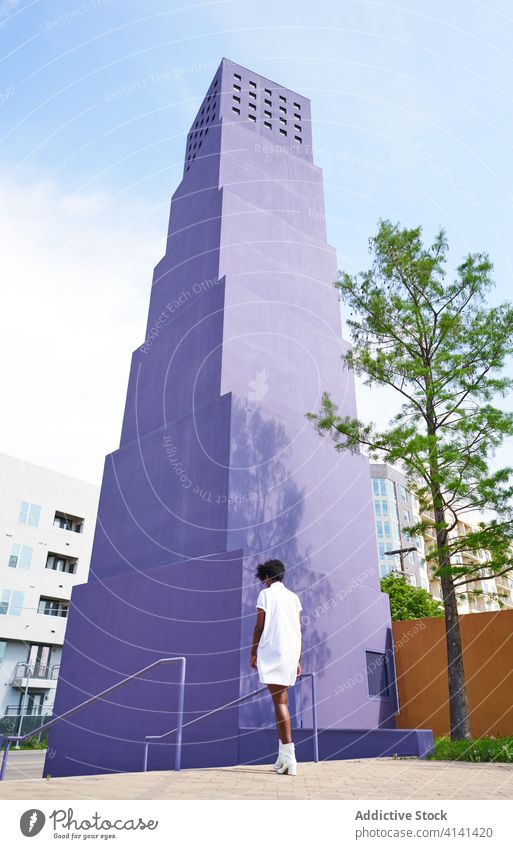 Stylish black woman walking on street against purple high rise building in downtown dress fashion trendy outfit female architecture style streetlamp urban