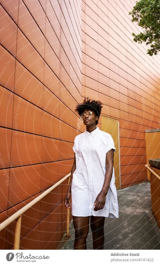 Confident African American woman in stylish outfit at entrance of contemporary building with orange tiled facade posing street fashion trendy exterior dress