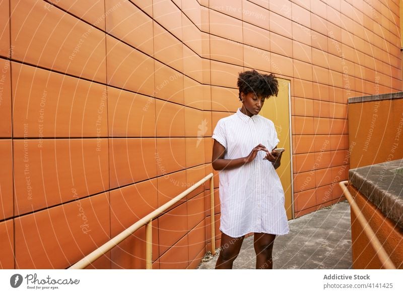 Focused black woman using smartphone on street against vivid exterior of modern building messaging pathway chill browsing smile social media facade surfing read