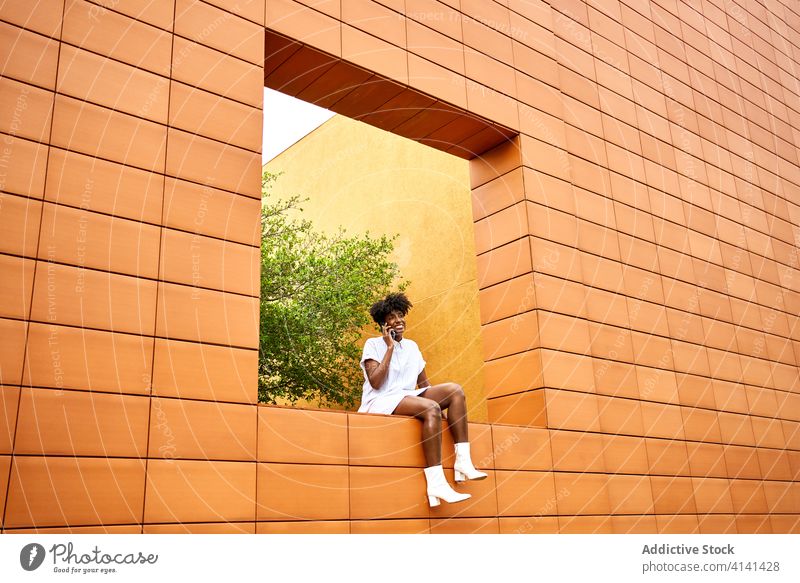 Stylish joyful black woman talking on phone on street near building with colorful exterior smartphone call architecture urban park chat modern conversation