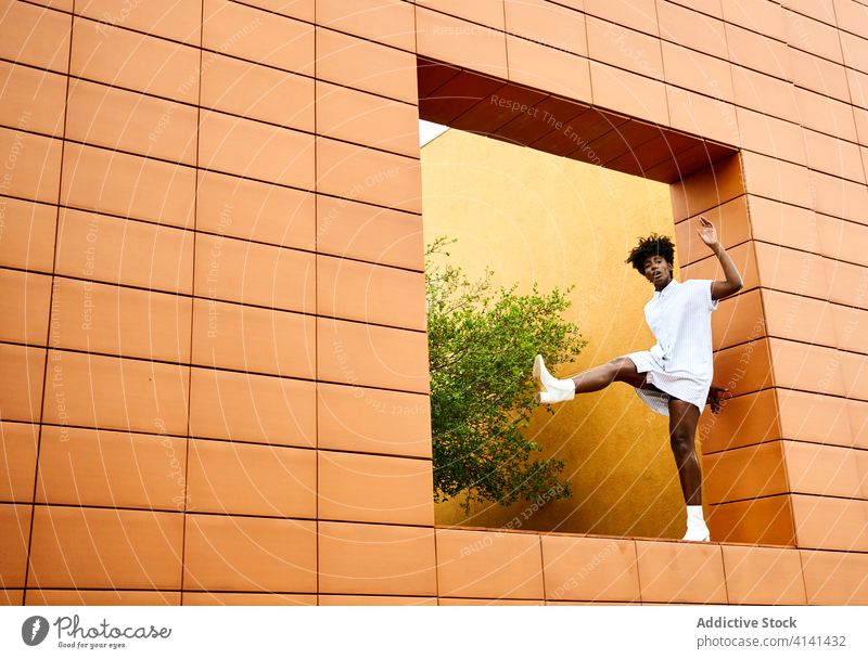 Carefree black woman in trendy clothes at window hole of contemporary building with orange tiled exterior architecture fashion carefree dress boot heel ethnic