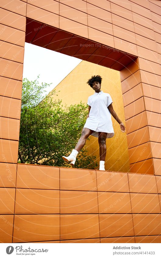 Carefree black woman in trendy clothes at window hole of contemporary building with orange tiled exterior architecture fashion carefree dress boot heel ethnic