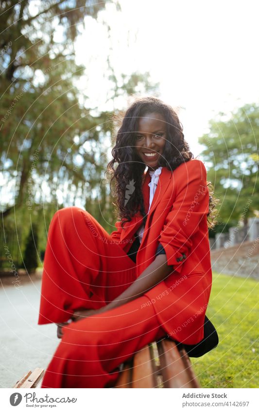 Cheerful black model in red outfit sitting on bench outdoors fashion appearance colorful cloth glamour elegant cheerful park woman vogue suit apparel slim
