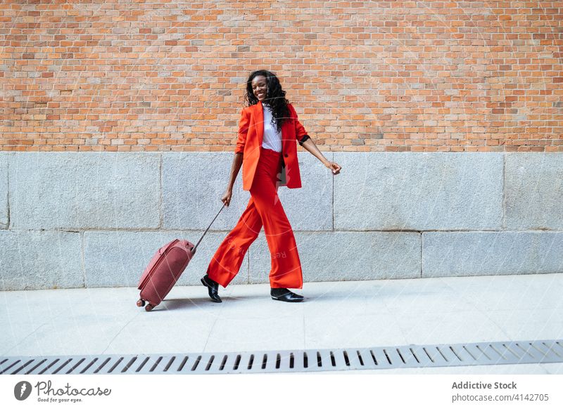 Black woman walking with suitcase on pavement fashion glamour style red brick wall building gesture elegant colorful appearance cloth stroll cheerful vogue