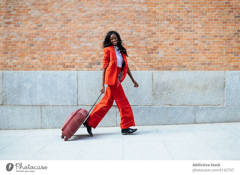 Black woman walking with suitcase on pavement fashion glamour style red brick wall building gesture elegant colorful appearance cloth stroll cheerful vogue