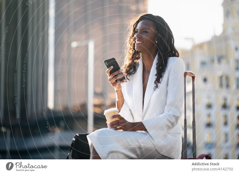 Smiling stylish black woman in white suit surfing smartphone businesswoman elegant browsing text messaging executive trip suitcase cellphone travel wait street