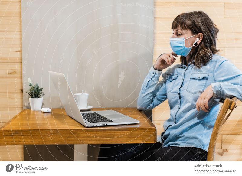 Concentrated woman working distantly on laptop in cafeteria freelance browsing earbuds focus medical mask coronavirus remote internet using female denim shirt