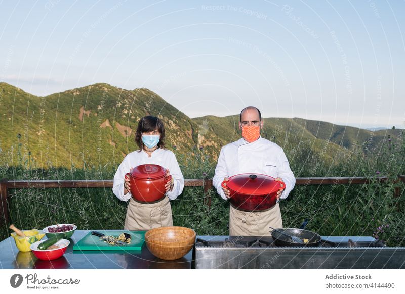 Chefs in aprons with prepared dishes in countryside man woman chef kitchen mountain terrain saucepan professional job rustic restaurant cook uniform mask