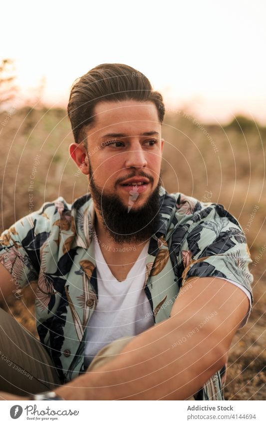 Pensive bearded man sitting on grass in field at sunset contemplate hipster mouth opened harmony idyllic countryside sky weekend hairstyle muscular nature