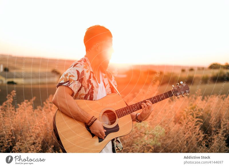 Male musician playing guitar in countryside at sunset hipster grass sky field harmony man musical instrument guitarist artificial light stand happy idyllic