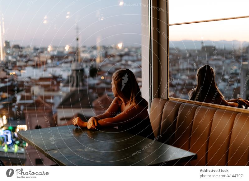 Young woman enjoying cityscape from cafe rooftop window evening madrid sunset cozy urban spain glass wall comfort relax restaurant reflection admire lounge