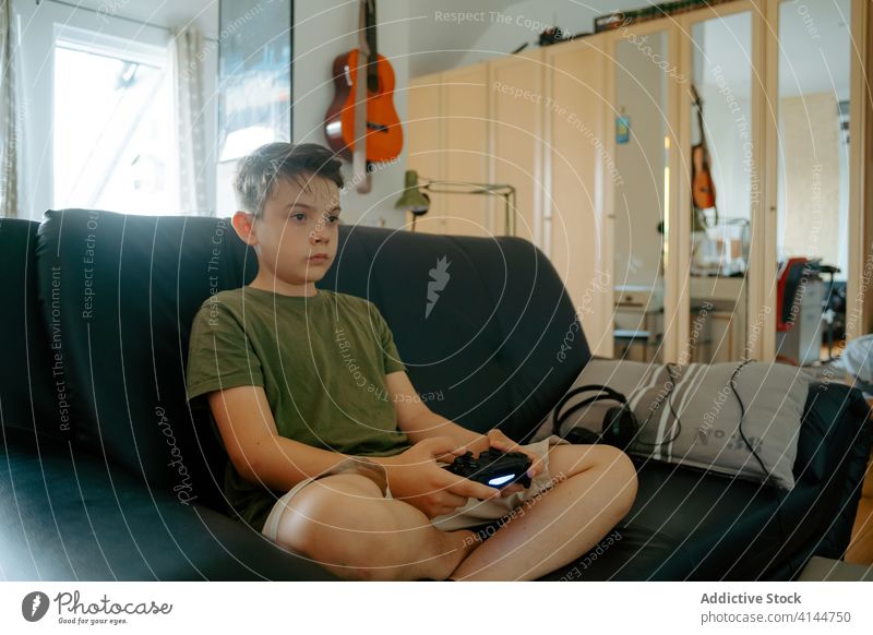 Focused child playing video game on couch boy joystick sofa legs crossed home entertain device gadget leisure concentrate kid little casual relax focus comfort
