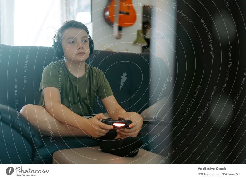 Focused child playing video game on couch boy joystick sofa legs crossed headphones home entertain device gadget leisure concentrate kid little casual relax